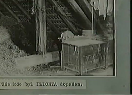 A period photograph of a bare attic under open rafters, a wooden chest beside a plank wall and washing hung from a beam, a typed Czech caption pasted beneath reading the attic where Plichta was caught