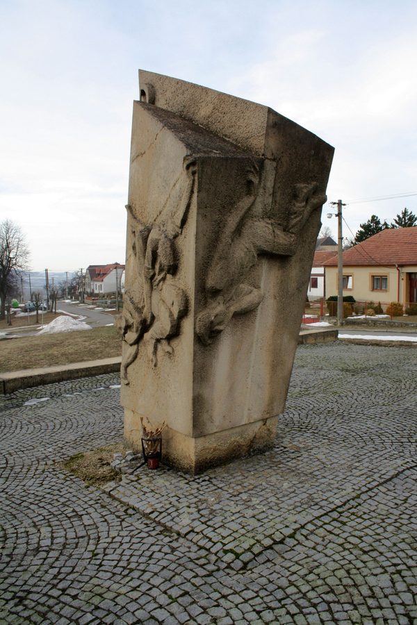 A tall stone monument on the cobbled village square in Babice, carved in relief with falling figures, a candle burning at its foot, houses and open country behind