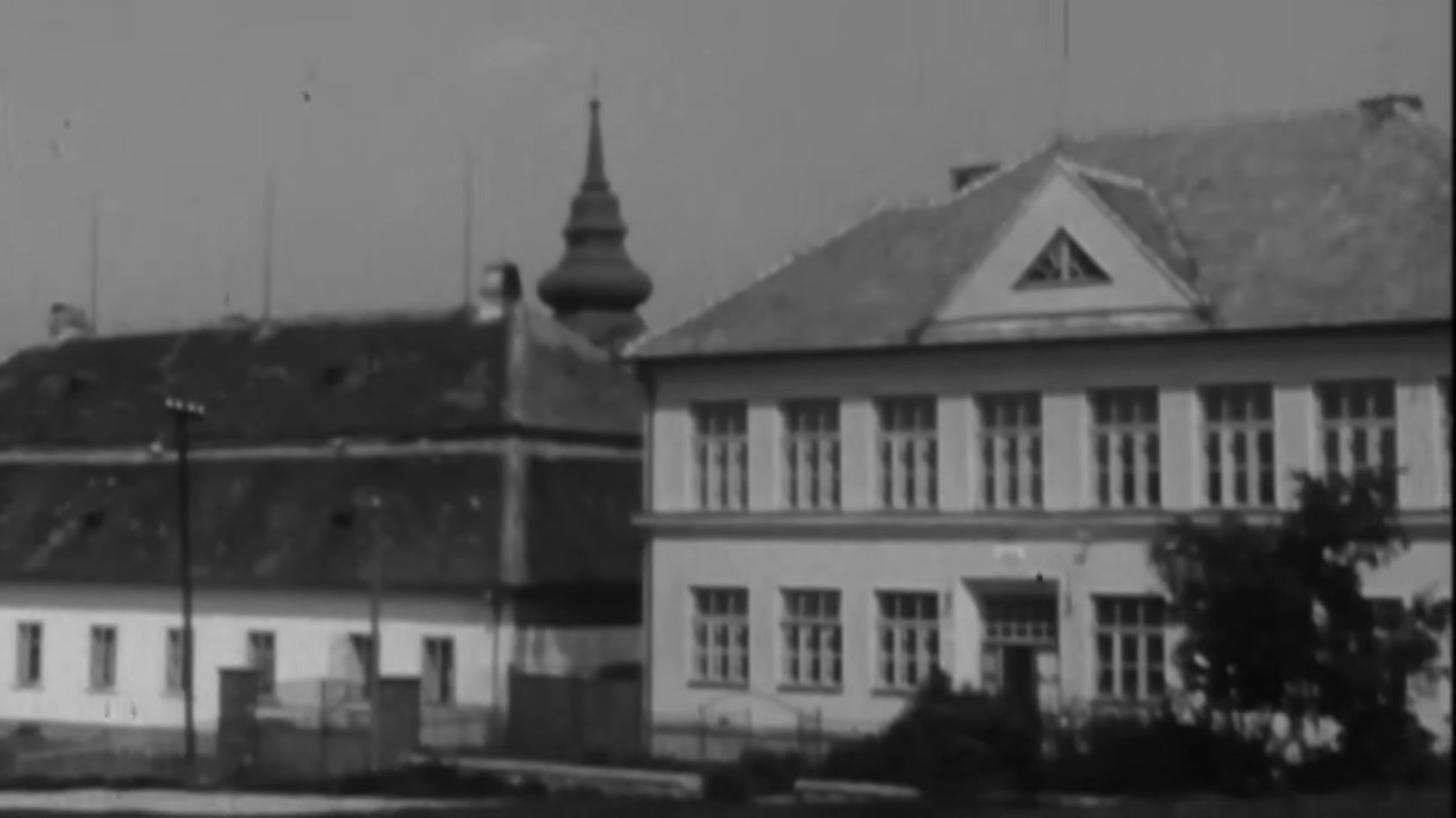 A period black and white photograph of the two storey village school in Babice, a long low building beside it and a church tower behind