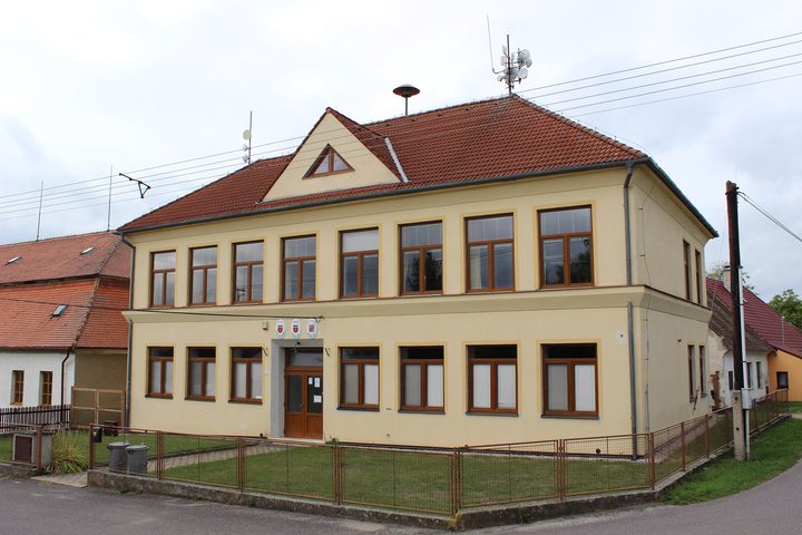 The former school building in Babice, two storeys, pale yellow with a red tiled roof, municipal crests and a loudspeaker above the door, photographed in the present day