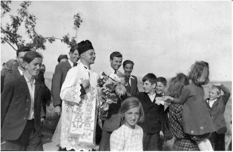 Jan Bula in a lace surplice and an embroidered stole, a bouquet in his hands, walking in the open among village children and men in Sunday clothes