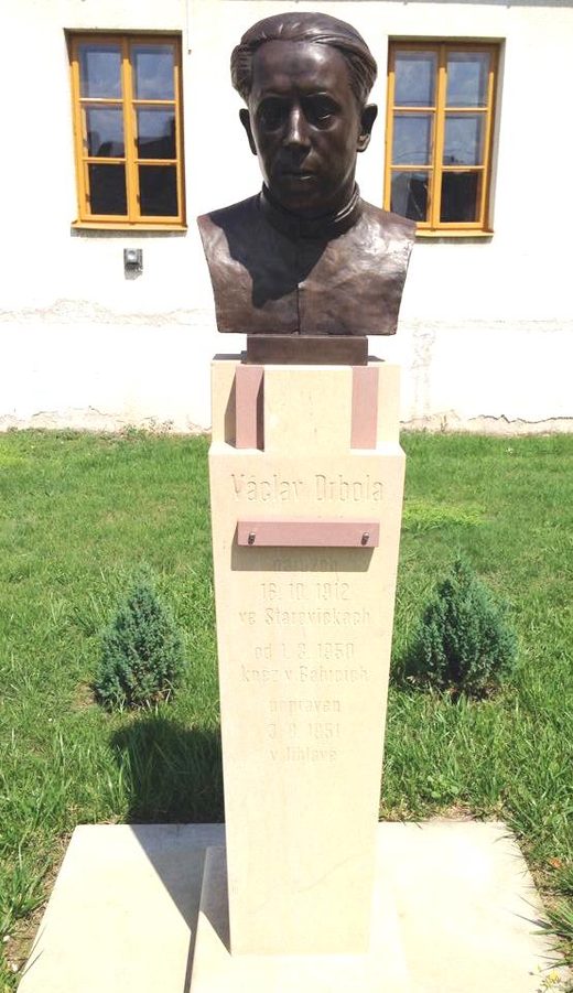 A bronze bust of Vaclav Drbola on a pale stone plinth in the grass before the parish house, the inscription giving his birth in 1912, his years as priest in Babice from 1950 and his execution in Jihlava in 1951