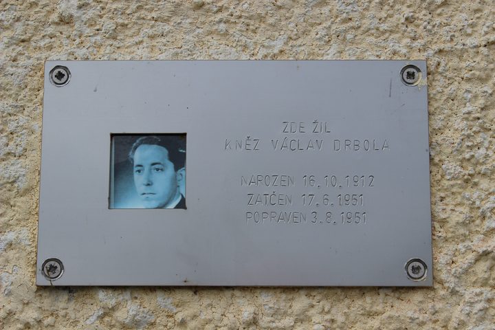 A metal plaque on a rendered wall, a small portrait set into it beside Czech lettering that reads here lived the priest Vaclav Drbola, born 16 October 1912, arrested 17 June 1951, executed 3 August 1951
