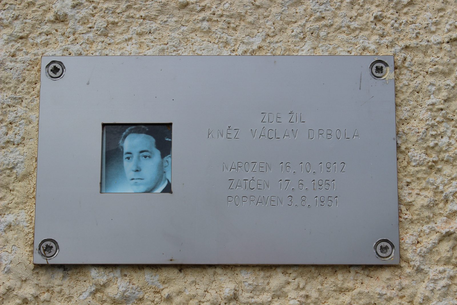 A metal plaque on a rendered wall, a small portrait set into it beside Czech lettering that reads here lived the priest Vaclav Drbola, born 16 October 1912, arrested 17 June 1951, executed 3 August 1951