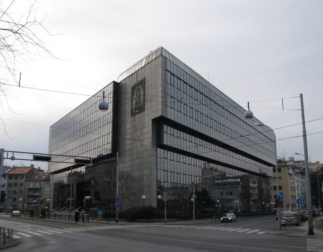 The INA Naftaplin building in Zagreb, a low modernist block of dark glass and stone on a street corner with tram wires overhead and a relief panel of a drilling rig on its face