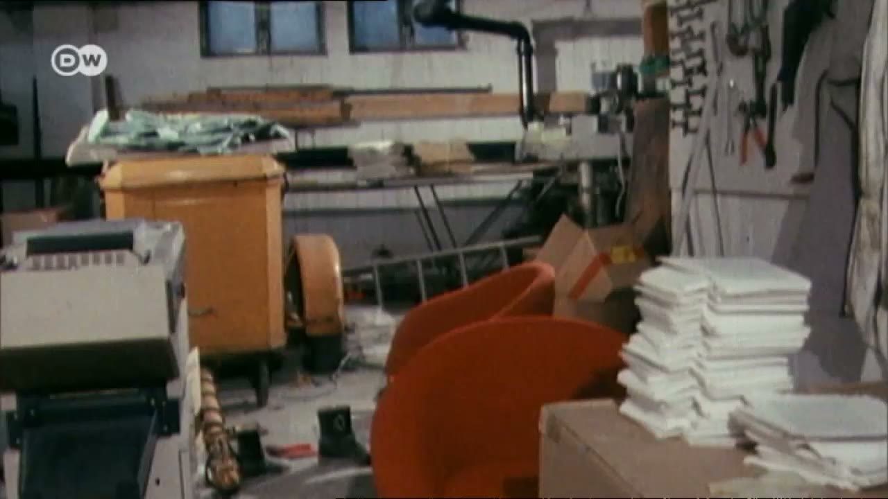 The inside of a small workshop: a printing machine on the left, an orange bin and a roll of paper, a workbench with a drill press and hand tools hanging on the wall, and stacks of white paper on a crate in the foreground