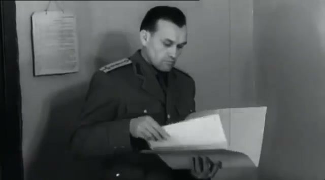 A Securitate officer in uniform, seated, reading an open file held on his knee, a printed notice pinned to the wall behind him
