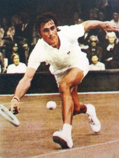 Ilie Năstase reaching low for a forehand on a grass court in white tennis clothes, spectators in the stand behind him