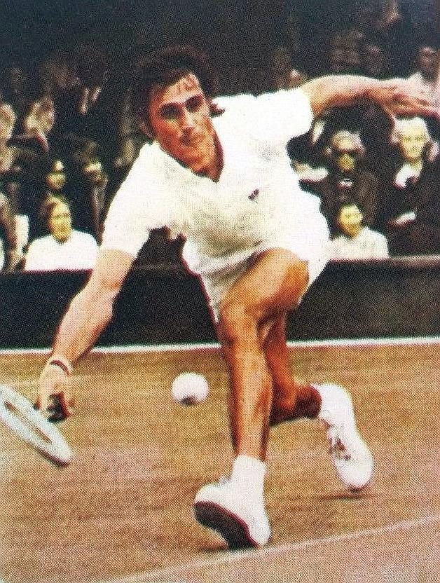 Ilie Năstase reaching low for a forehand on a grass court in white tennis clothes, spectators in the stand behind him