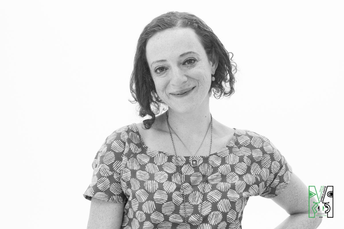 A black and white portrait of Irene Lusztig smiling, one hand on her hip, against a white background, with the Ambulante documentary festival logo in the lower corner