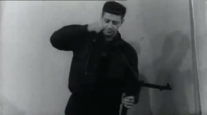 A man in a dark jacket pulls a garment over his head, a submachine gun in his other hand, standing against a bare wall
