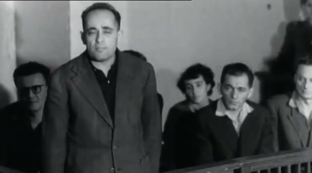 A man stands at the front of a wooden dock, hands at his sides, with five others seated behind him, one of them a woman and one wearing glasses, filmed in black and white