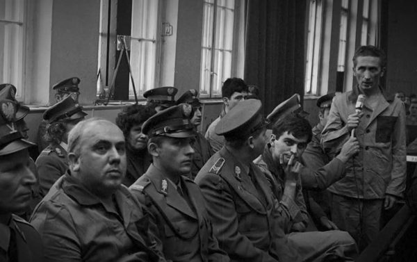Zdzislaw Marchwicki standing with a hand-held microphone at the trial, militia officers in uniform seated in front of him
