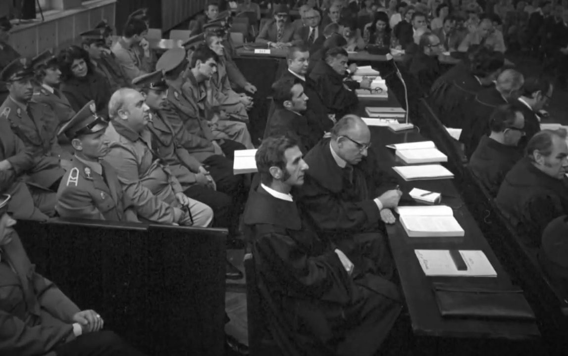 The trial hall: advocates in robes at a long table with their files, the defendants and their militia escorts on the benches beside them, the public gallery behind