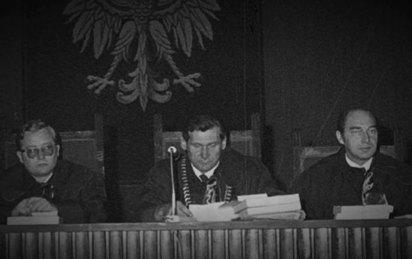 Three men in judges' robes at a long bench, papers in front of them, a large Polish eagle on the wall above