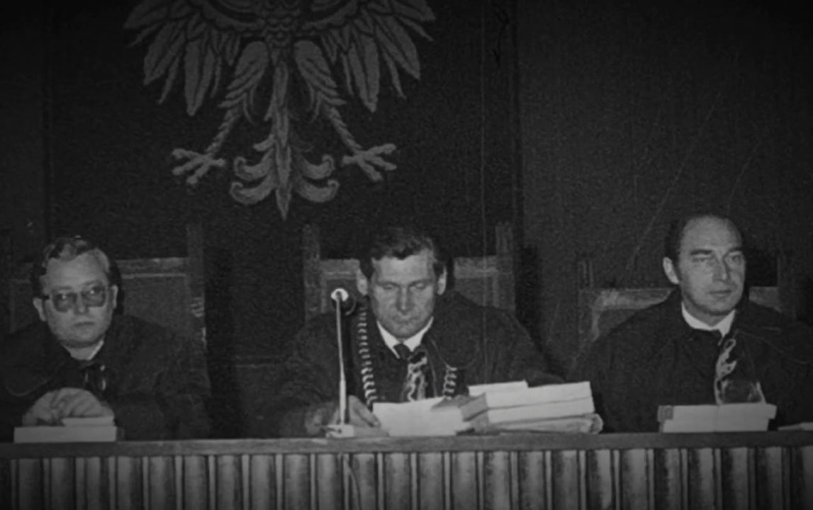 Three men in judges' robes at a long bench, papers in front of them, a large Polish eagle on the wall above