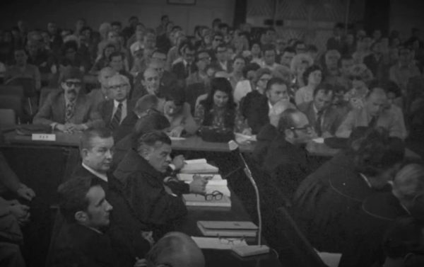 A packed hall of spectators rising behind the prosecution tables, advocates in robes in the foreground