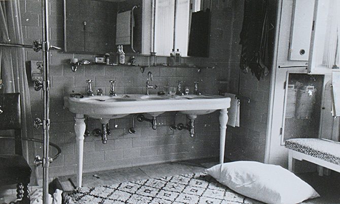 The bathroom of Jan Masaryk's flat on 10 March 1948: a double washbasin on a stand with turned legs, a mirror and bottles on the shelf above it, a shower fitting and a chair at the left, and a pillow lying on the floor beside the patterned mat