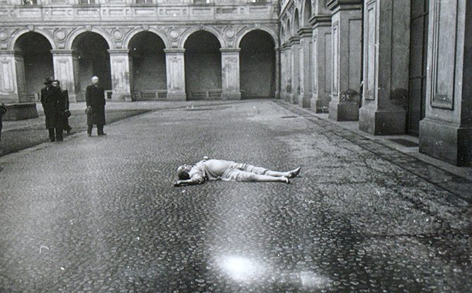 The paved courtyard of the Czernin Palace on 10 March 1948 seen from the far side: the body lying on the cobbles in the middle of the yard, the arcade of the wing behind it and three men standing at the far left