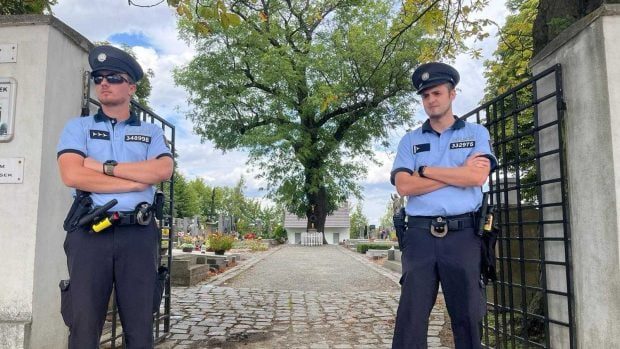 Two uniformed Czech police officers standing with folded arms at the open iron gate of the village cemetery at Lány on 2 September 2026, the graves and a large tree behind them