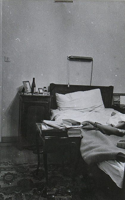 The bedroom of Jan Masaryk's flat on 10 March 1948: the unmade bed with a reading lamp on a bracket above it, a night table carrying a bottle, a glass and a framed picture, and a small table at the bedside piled with books and papers