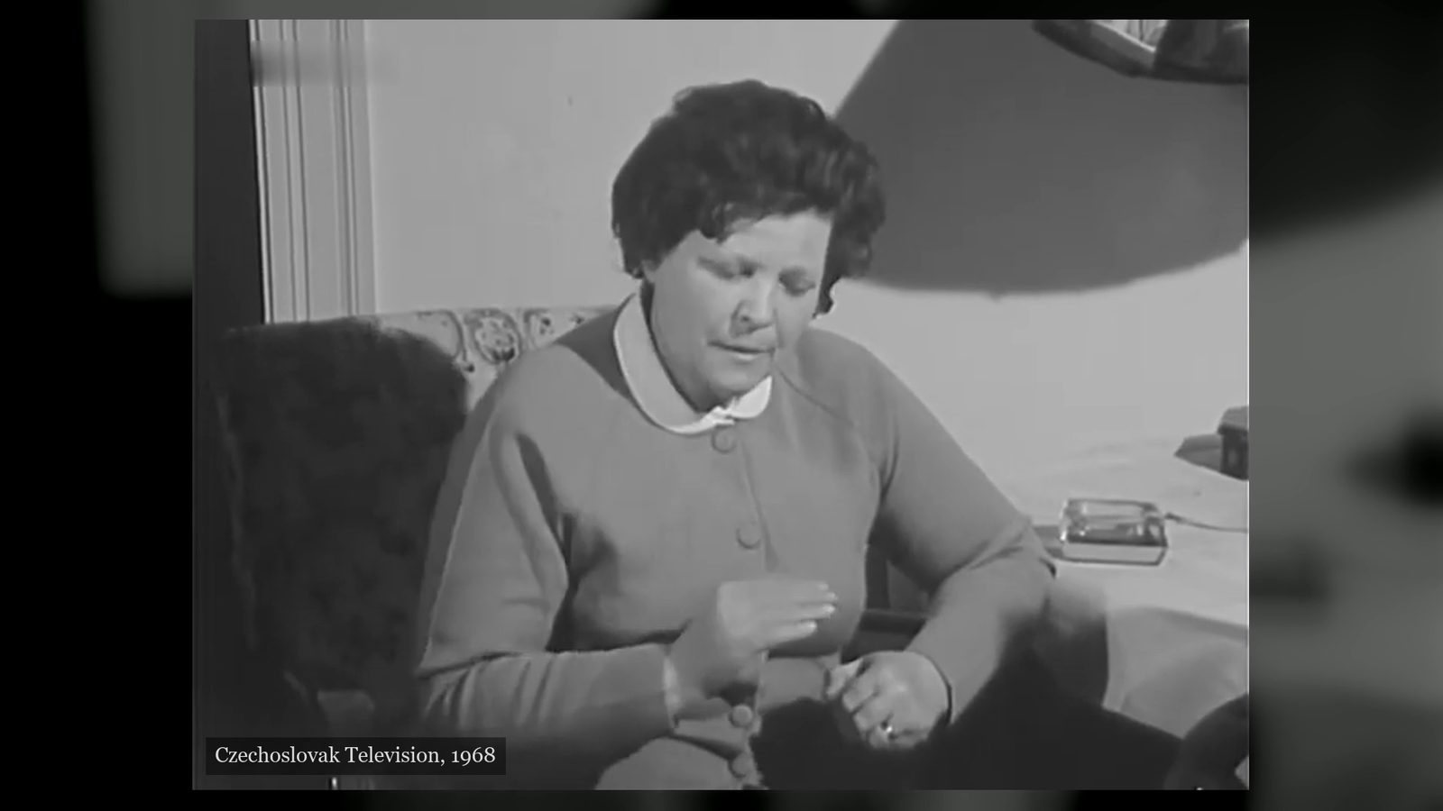 Marie Topinková seated on a sofa in a flat, in a buttoned cardigan and a white collar, her hands raised in front of her as she speaks, filmed by Czechoslovak Television in 1968