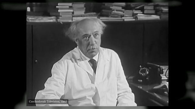 Professor Kácl, white haired and in a white coat, seated in an office with stacks of journals on the cupboard behind him and a telephone on the desk at his side, filmed by Czechoslovak Television in 1968