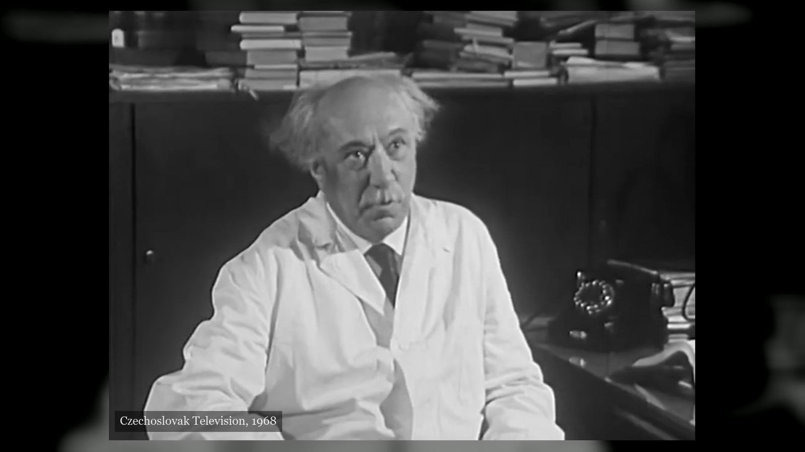 Professor Kácl, white haired and in a white coat, seated in an office with stacks of journals on the cupboard behind him and a telephone on the desk at his side, filmed by Czechoslovak Television in 1968