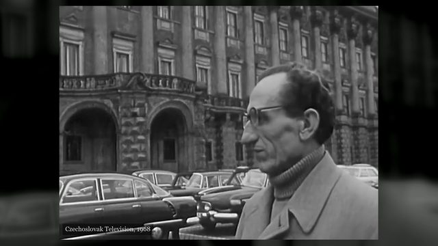 A man in a raincoat and glasses, seen in profile, standing on Loreta Square with the long facade of the Czernin Palace and a row of parked cars behind him, filmed by Czechoslovak Television in 1968