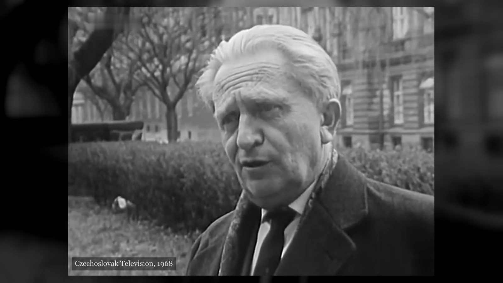 Jaromír Tesař, white haired, in a dark coat and tie, standing in a city park with bare trees and a large building behind him, filmed by Czechoslovak Television in 1968