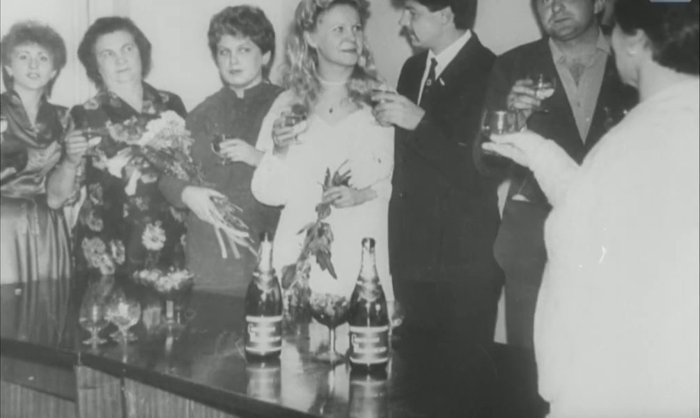 A gathering indoors: men and women standing with raised glasses and bouquets, two bottles on the table in front of them, a moustached man in a dark suit among them