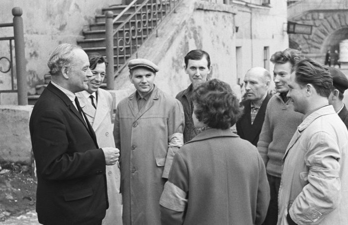 A group of men standing in a courtyard in coats and jackets, several with armbands on their left sleeves, listening to an older man in a dark suit