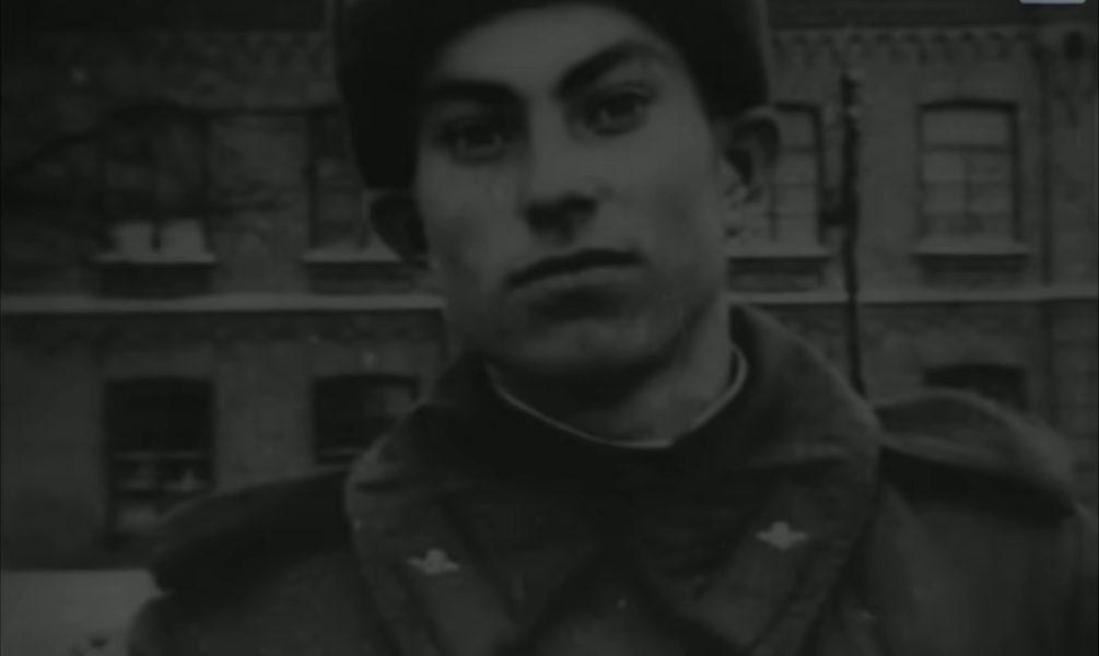 A young man in a fur cap and a heavy army greatcoat standing in the snow outside a brick building, looking into the camera