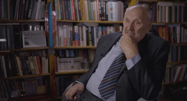 An elderly man in a dark suit and striped tie sitting in front of full bookshelves, one hand raised to his chin