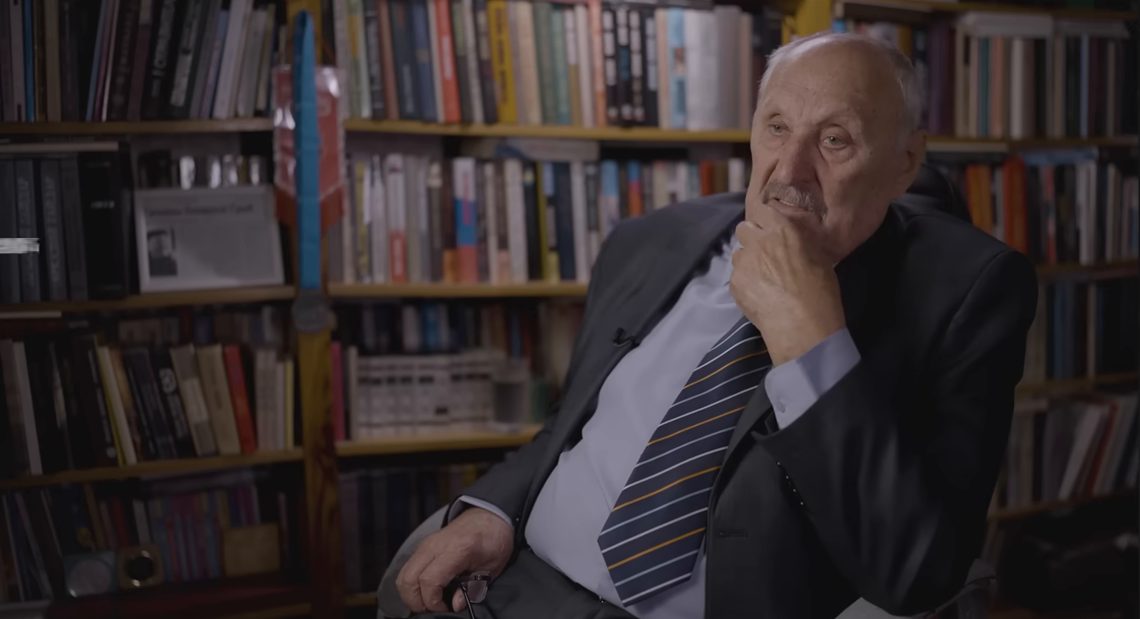 An elderly man in a dark suit and striped tie sitting in front of full bookshelves, one hand raised to his chin