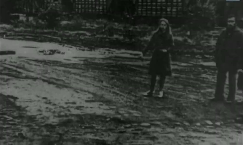 A rutted village track in poor weather: a woman in a headscarf and dark coat standing in the middle of it, a man waiting at the right edge of the frame