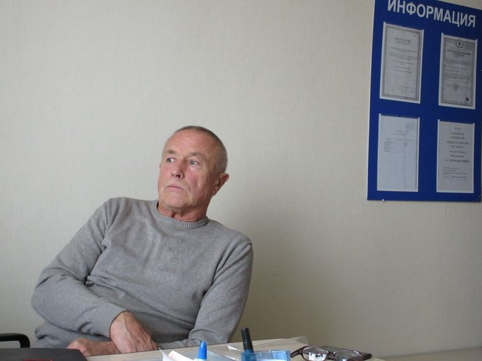 An elderly man in a grey jumper sitting at a table in a plain office, looking away from the camera, an information board on the wall behind him