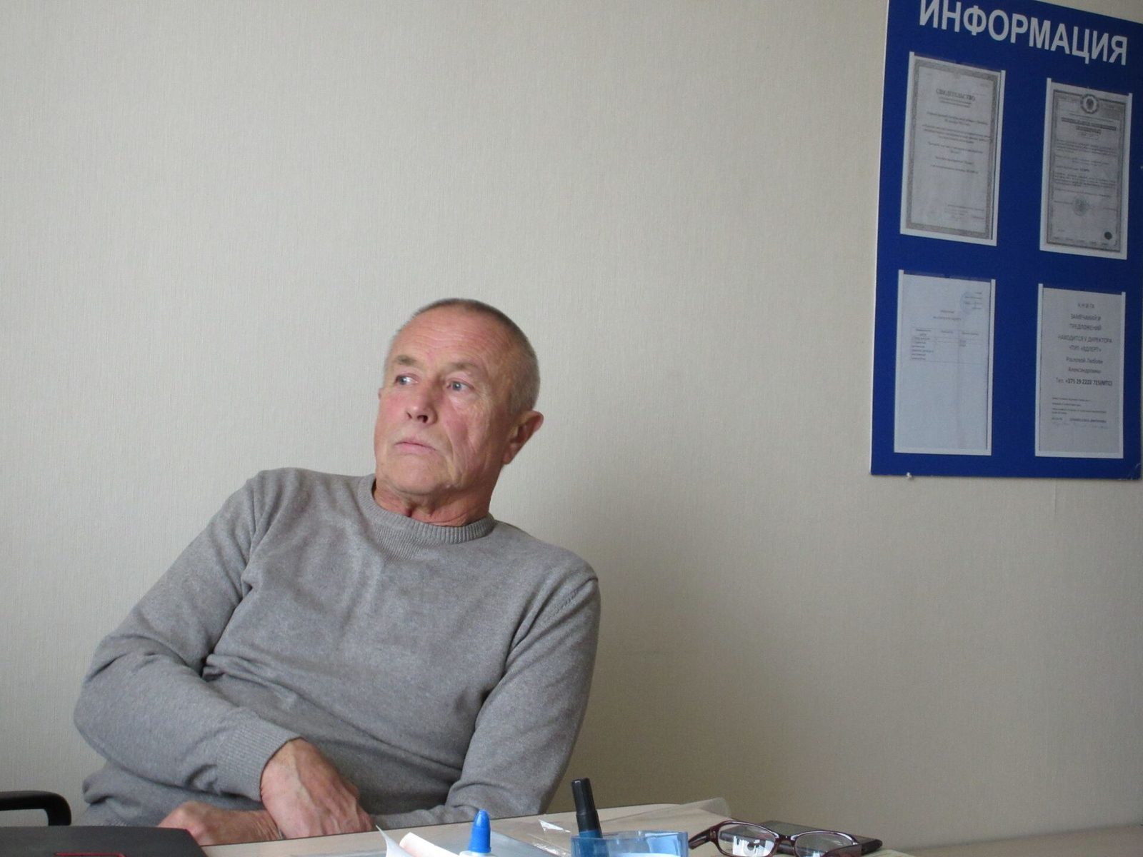 An elderly man in a grey jumper sitting at a table in a plain office, looking away from the camera, an information board on the wall behind him