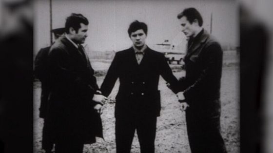 Three men standing on open ground: a young man in a dark buttoned coat in the centre, flanked by two others in coats, a vehicle parked behind them