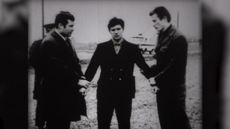 Three men standing on open ground: a young man in a dark buttoned coat in the centre, flanked by two others in coats, a vehicle parked behind them