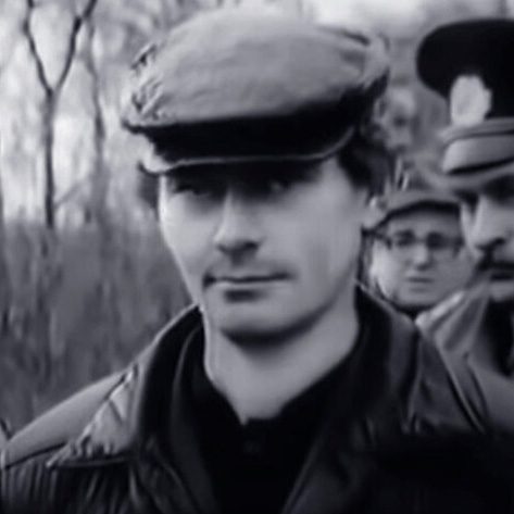 Gennady Mikhasevich in a flat cap and a dark jacket, photographed outdoors among bare trees after his detention, with uniformed officers behind him