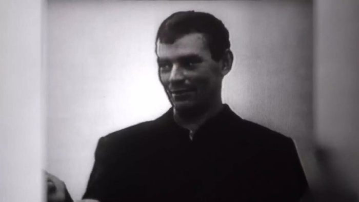 A man in a dark shirt standing against a plain wall in a bare room, half smiling, filmed in black and white during questioning
