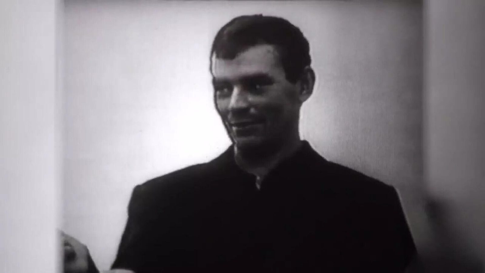 A man in a dark shirt standing against a plain wall in a bare room, half smiling, filmed in black and white during questioning