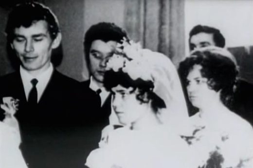 A wedding photograph in black and white: a groom in a dark suit at the left, a bride in a veil and headdress in front of him, guests standing behind