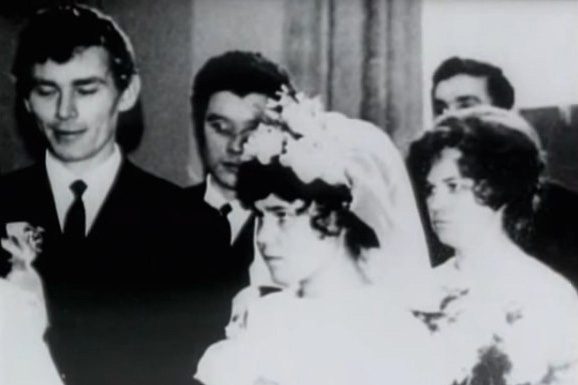 A wedding photograph in black and white: a groom in a dark suit at the left, a bride in a veil and headdress in front of him, guests standing behind