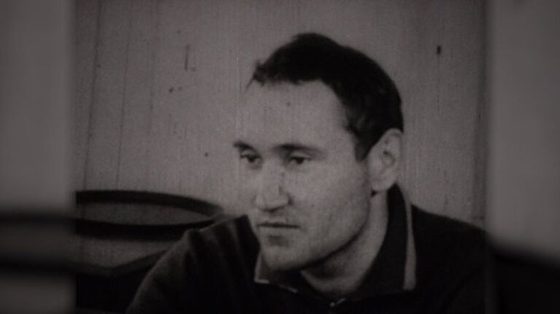 A young man in a dark jacket sitting in a bare room and looking down, photographed during a confrontation with another witness
