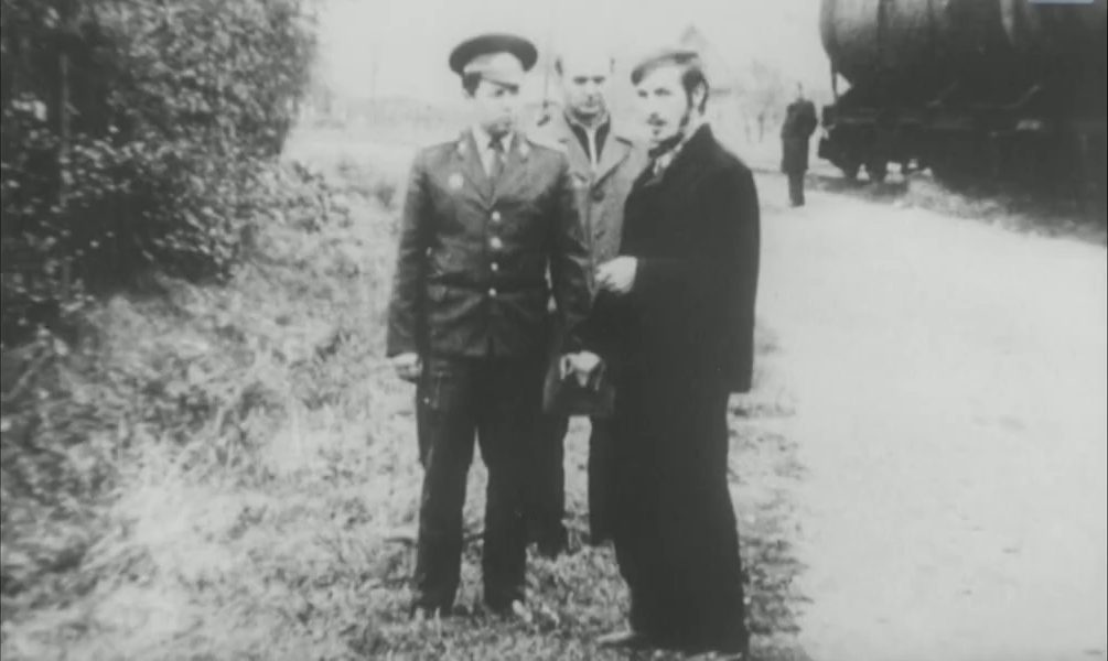 A roadside verge with a rail wagon behind it: a militiaman in uniform and peaked cap, a second man in a jacket, and a dark haired man in a long coat standing between them