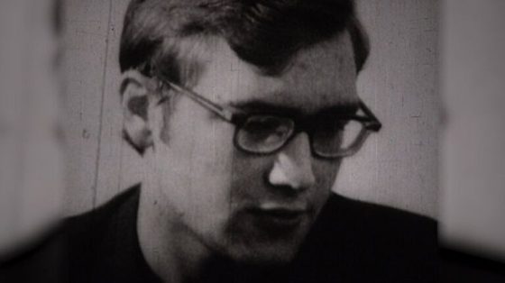 A young man in dark rimmed glasses, head lowered, photographed in a bare room during questioning