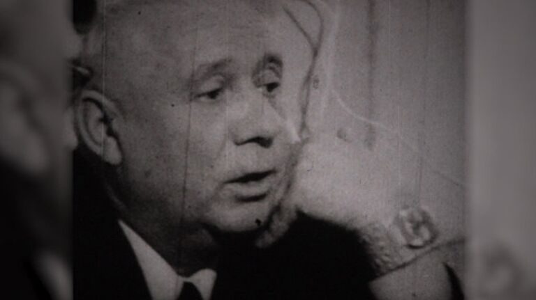 An elderly man in a dark suit and tie, mouth open in mid sentence, filmed close up against a pale wall