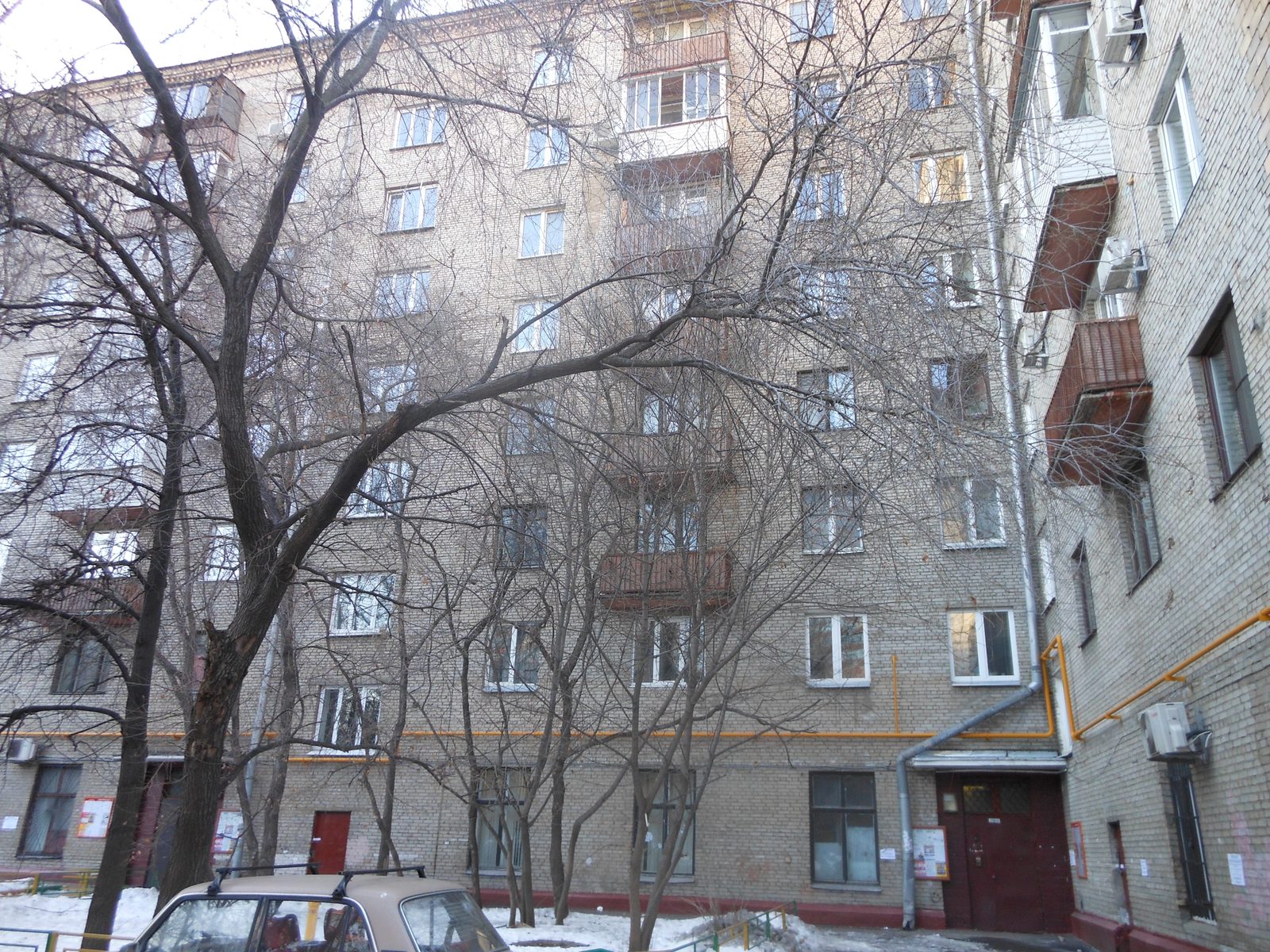 The courtyard side of a long brick apartment block in Moscow in winter: six storeys of windows and small balconies, bare trees in front, snow on the ground and a car parked at the foot of the building